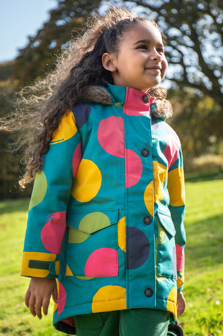 Girl outside wearing Frugi Penhale Parka in turquoise with green and yellow spots with a furry hood - made from PU-coated organic cotton, waterproof 3000HH, to represent Frugi Outerwear, sold at Babipur.