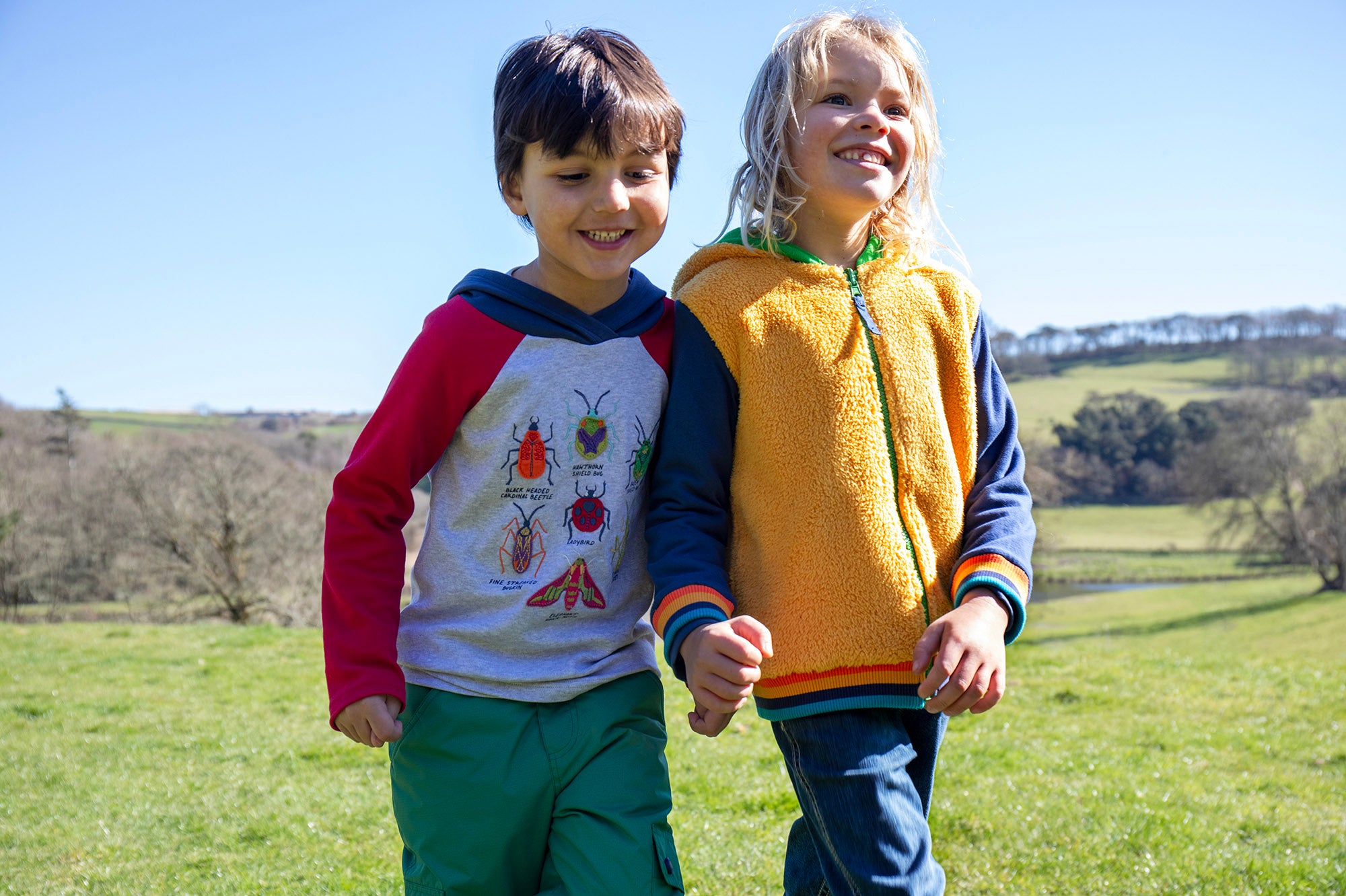 A child standing outside and wearing the Frugi compass kids hooded top in grey bug next to another child wearing the helston reversible hoodie available at Babipur.