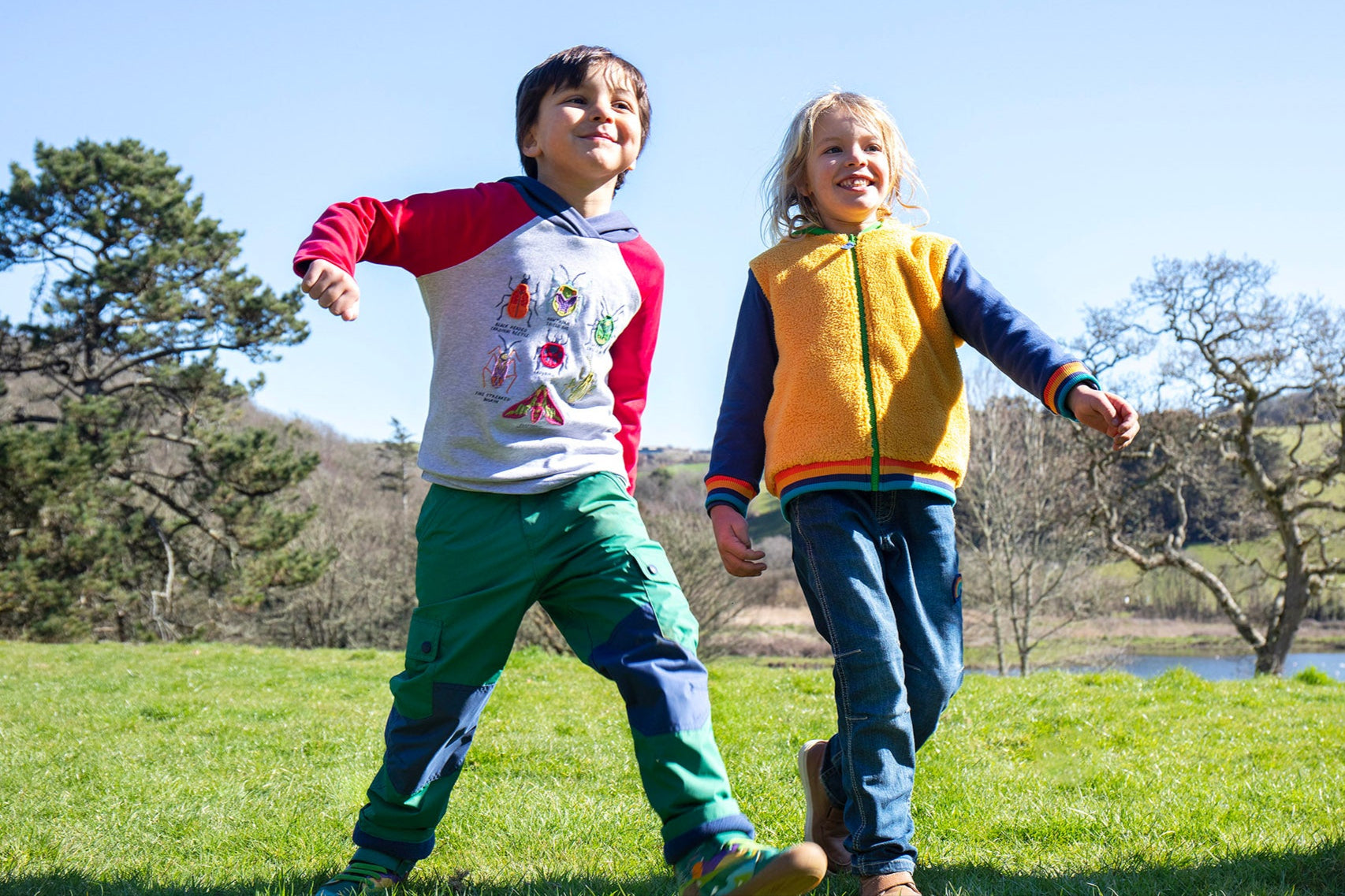 A child walking on grass and wearing the Frugi compass kids hooded top in grey bug next to another child wearing the helston reversible hoodie available at Babipur.
