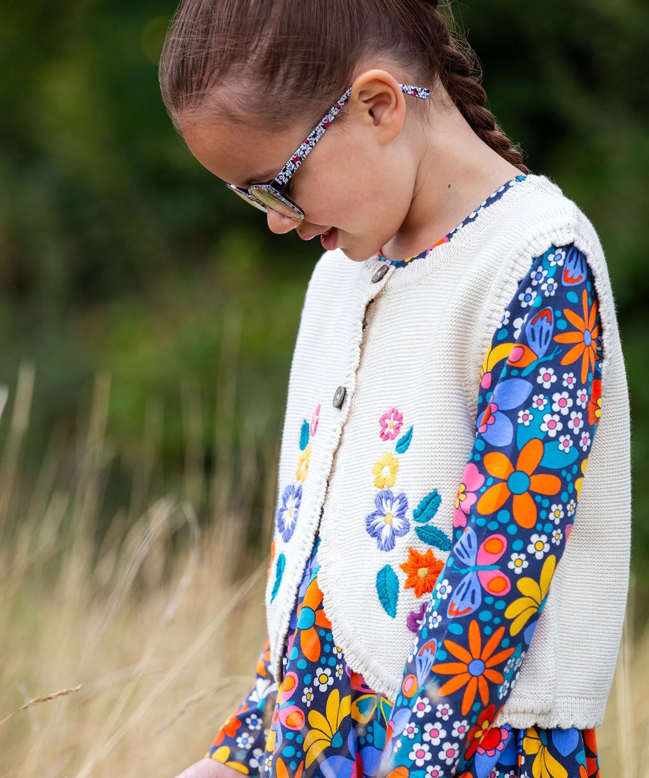 A close up of a girl wearing the Frugi Oatmeal Flowers Connie Cotton Knitted Sleeveless Cardigan available at Babipur showing  the embroidery detail. 