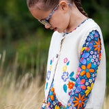 A close up of a girl wearing the Frugi Oatmeal Flowers Connie Cotton Knitted Sleeveless Cardigan available at Babipur showing  the embroidery detail. 