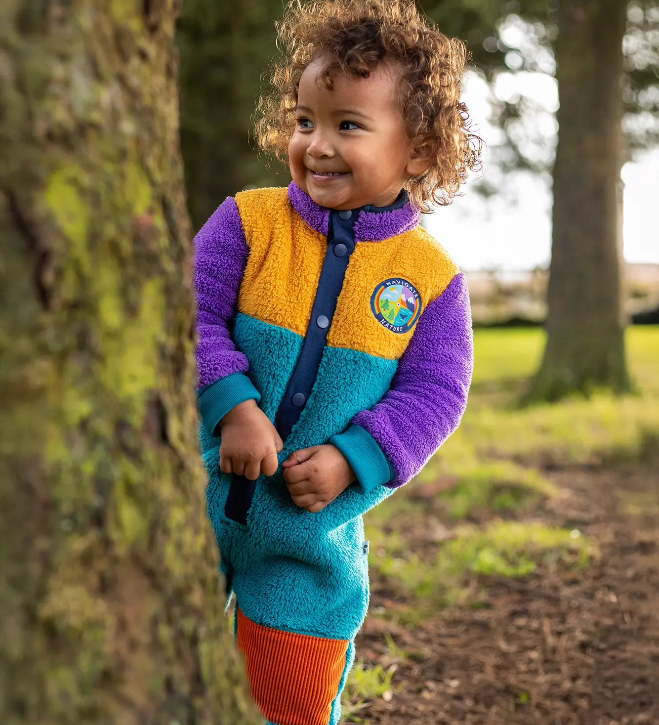Frugi Cord Fleece Snuggle Suit - Hotchpotch worn by child - a cosy blue, purple and yellow all-in-one with orange cord knee patches.