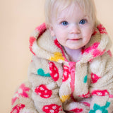 A toddler sitting down and wearing the Frugi kids cosy ted snuggle suit in mushroom field available at Babipur. A thick all in one hooded suit in beige with a cute mushroom and flower design. Features a full length zip in pink and matching pink cuffs