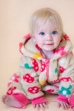 A toddler sitting down and wearing the Frugi kids cosy ted snuggle suit in mushroom field available at Babipur. A thick all in one hooded suit in beige with a cute mushroom and flower design. Features a full length zip in pink and matching pink cuffs