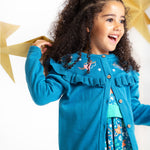 A happy child playing with gold, paper stars, wearing the Frugi Romi Ruffle Cardigan - Deep Water/Fairies. The cardigan is open showing a dress underneath