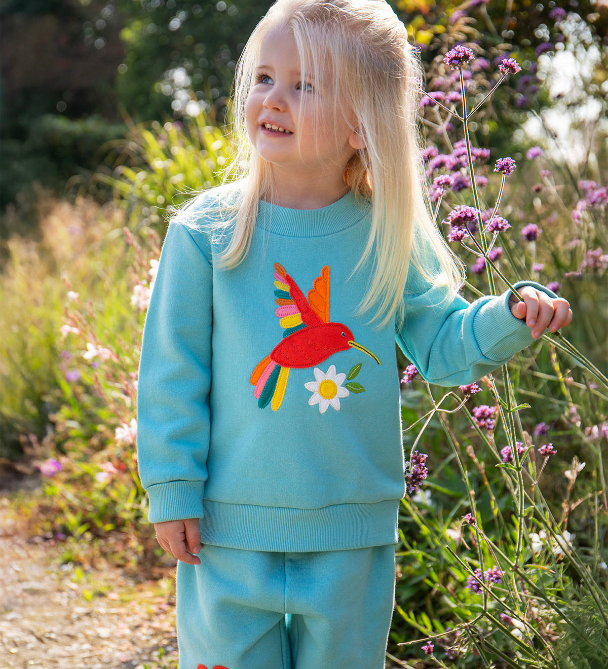 A child standing outdoors with purple flowers and grass in the background. wearing a light blue jumper with a playful red hummingbird design on the front and matching light blue joggers