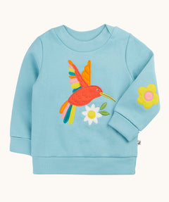Frugi organic cotton jumper laid flat. In light blue with a red playful hummingbird design on the front and a yellow flower patch on the sleeve.