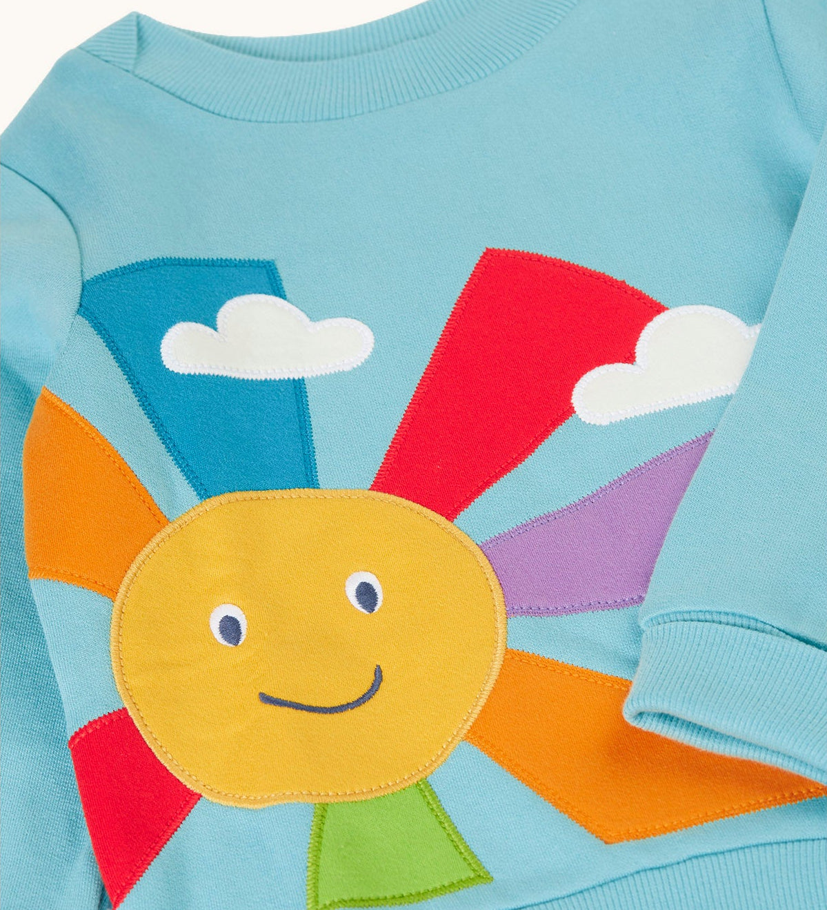 A close up of a light blue Frugi organic cotton jumper laid flat showing the smiling sun design on the front