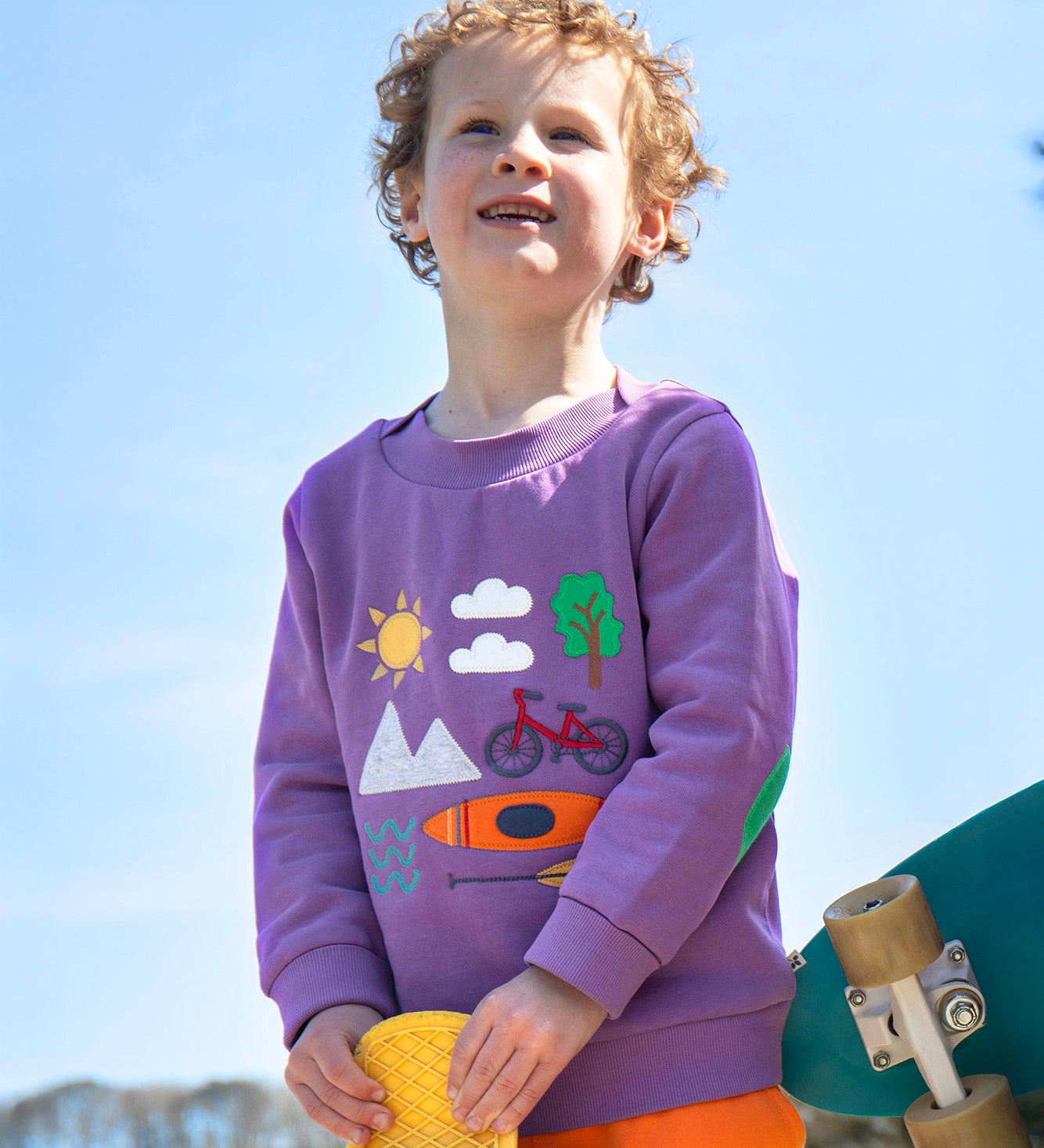 A smiling child standing outside and holding a yellow skateboard. Wearing the light purple jumper with various outdoor themed designs on the front