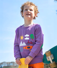 A smiling child standing outside and holding a yellow skateboard. Wearing the light purple jumper with various outdoor themed designs on the front