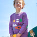 A smiling child standing outside and holding a yellow skateboard. Wearing the light purple jumper with various outdoor themed designs on the front