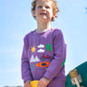 A smiling child standing outside and holding a yellow skateboard. Wearing the light purple jumper with various outdoor themed designs on the front