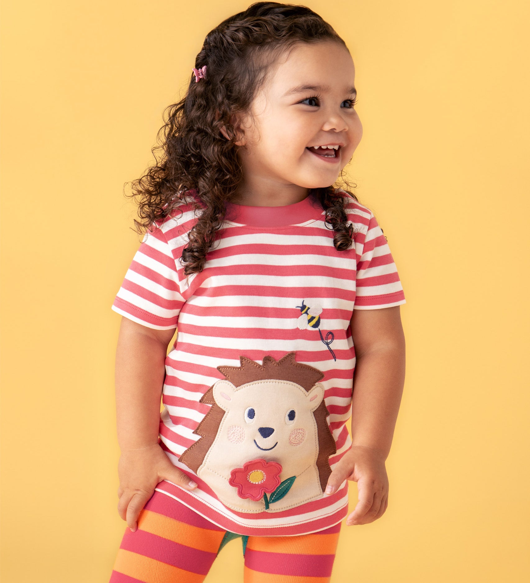 A smiling child standing in front of a light orange background. Wearing the Frugi white and pink stripe cotton short sleeve t-shirt with a cute hedgehog patch on the bottom.