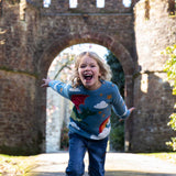 A child wearing the Frugi elwood dragon skies knitted jumper. A GOTS organic cotton jumper in light blue with a playful flying dragon design wrapping around the jumper. Features button fasteners on the shoulder. This children's knitted jumper is apart of a wide range of organic cotton clothing for children available here at Babipur. 