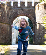 A child wearing the Frugi elwood dragon skies knitted jumper. A GOTS organic cotton jumper in light blue with a playful flying dragon design wrapping around the jumper. Features button fasteners on the shoulder. This children's knitted jumper is apart of a wide range of organic cotton clothing for children available here at Babipur. 