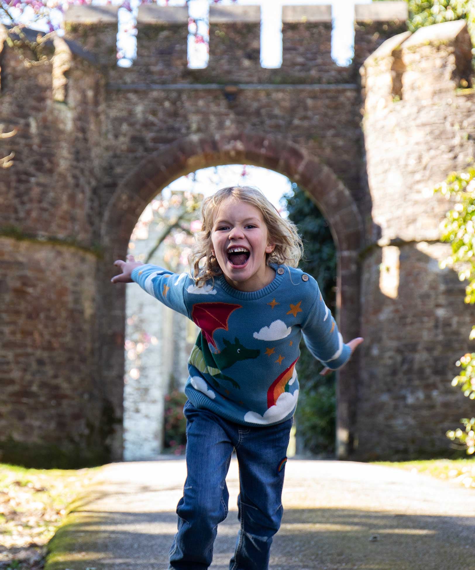 A child wearing the Frugi elwood dragon skies knitted jumper. A GOTS organic cotton jumper in light blue with a playful flying dragon design wrapping around the jumper. Features button fasteners on the shoulder. This children's knitted jumper is apart of a wide range of organic cotton clothing for children available here at Babipur. 