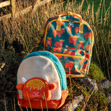 Frugi Highland cow character kids backpack standing next to the explorer  style backpack on a grassy mount
