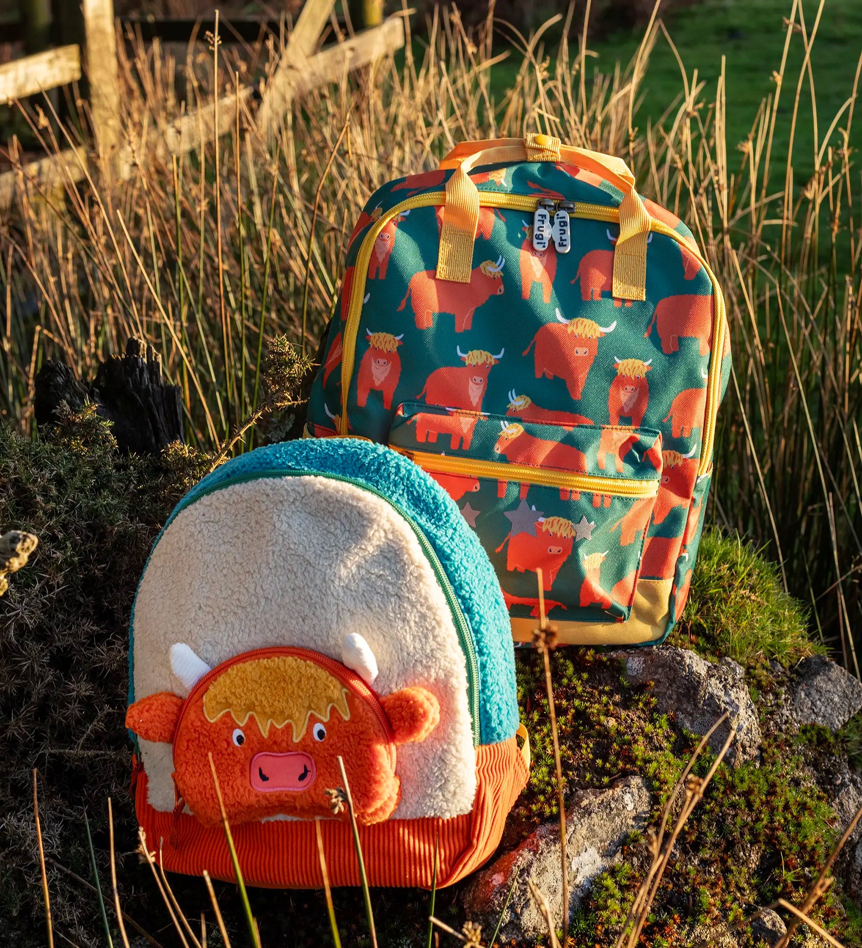 Frugi Highland cow character kids backpack standing next to the explorer  style backpack on a grassy mount
