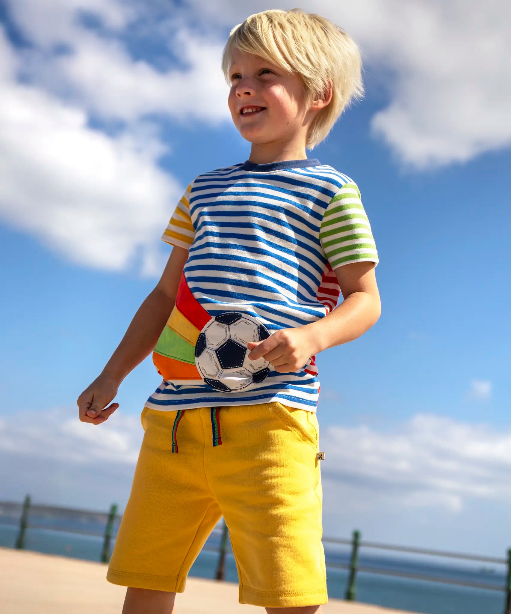 Child wearing Frugi's striped hotchpotch circular t-shirt with a football patch on the front and standing on the promenade