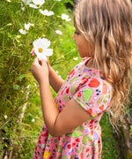 Child wearing Frugi's pink skater dress with repeating fruit and veg print and holding a white flower in a garden