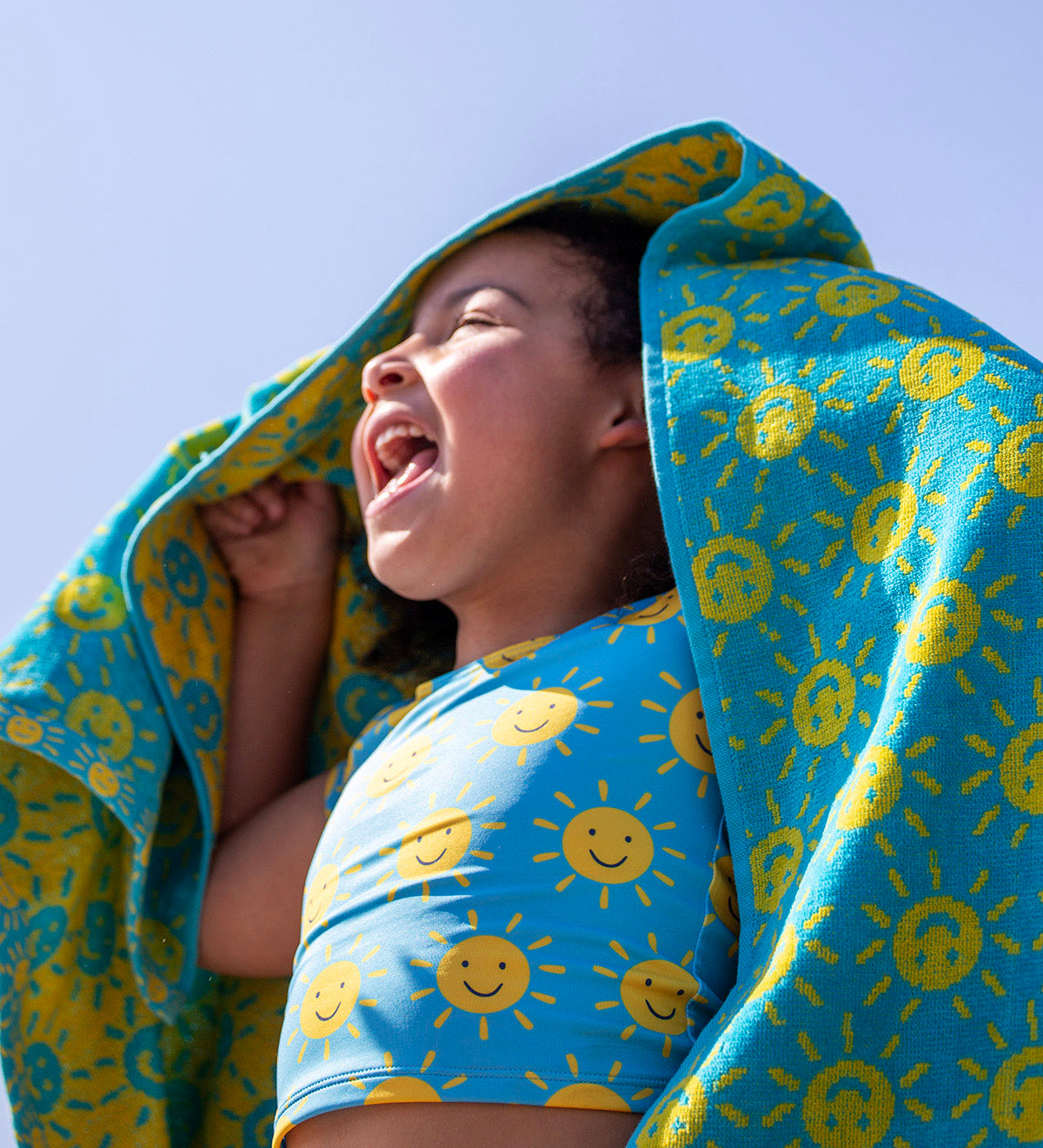 A child smiling with blue sky in the background and holding the turquoise Frugi hooded towel with a repeating smiling sun print. 