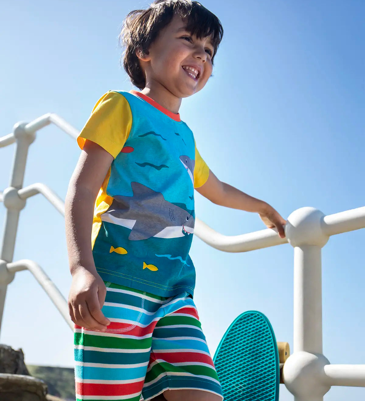 Child wearing Frugi's Iori hotchpotch swimming sharks design with yellow short sleeves t-shirt on concrete stairs