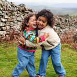 Girls hugging wearing Frugi, Juana Fleece jacket in cream with Alpine Blooms corduroy piping and mouse fairisle cardigans with blue jeans and brown boots.