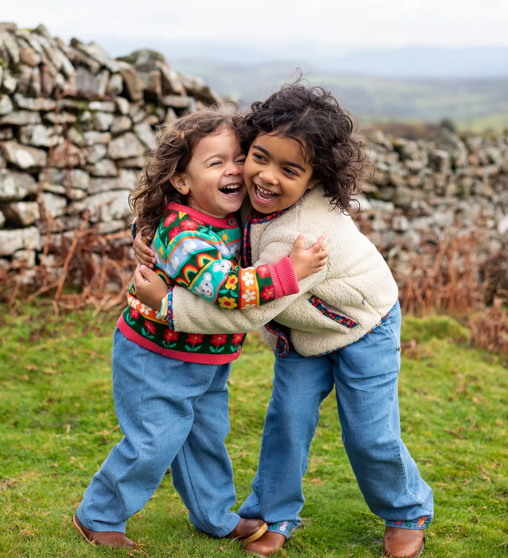 Girls hugging wearing Frugi, Juana Fleece jacket in cream with Alpine Blooms corduroy piping and mouse fairisle cardigans with blue jeans and brown boots.