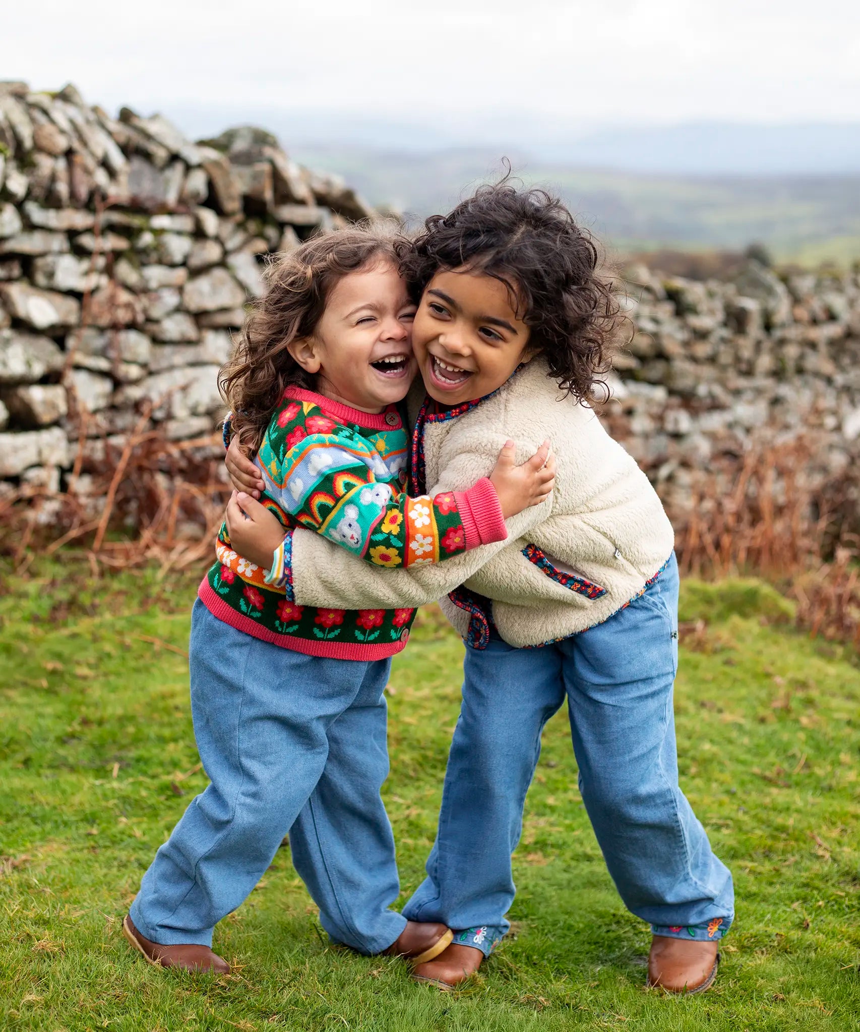 Girls hugging wearing Frugi, Juana Fleece jacket in cream with Alpine Blooms corduroy piping and mouse fairisle cardigans with blue jeans and brown boots.