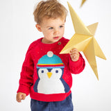 A child playing with a gold, paper star, wearing the Caleb Character Knit Jumper - True Red/Penguin. The jumper has a large penguin face and body, and the penguin is wearing a light blue hat