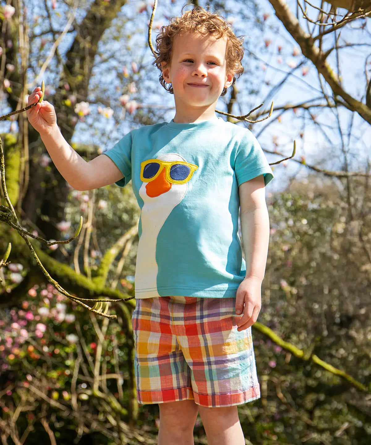 Child wearing Frugi 100% organic cotton Archie slub short in a pastel check design with a blue t-shirt showing trees in the background.