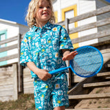 Child wearing Frugi's Kai shorts in blue with repeating beach inspired designs and holding a tennis racket 