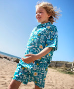 Child wearing Frugi's Kai shorts in blue with repeating beach inspired designs and walking on sand
