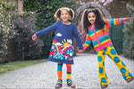 A child wearing the Frugi Kari navy rainbow dress and standing next to another child wearing the Frugi bight rainbow striped cardigan and super dot trousers available at Babipur.