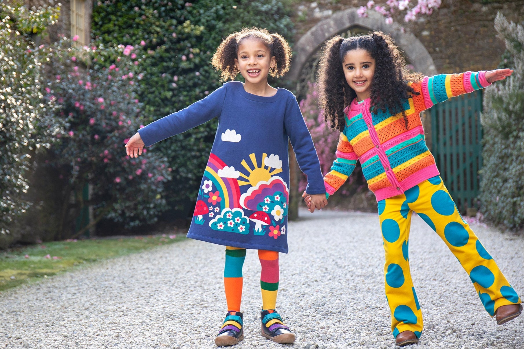 A child wearing the Frugi Kari navy rainbow dress and standing next to another child wearing the Frugi bight rainbow striped cardigan and super dot trousers available at Babipur.