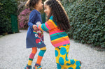 A child wearing Frugi Kari navy rainbow dress and playing with another child wearing the Frugi bight rainbow striped cardigan and super dot trousers available at Babipur.