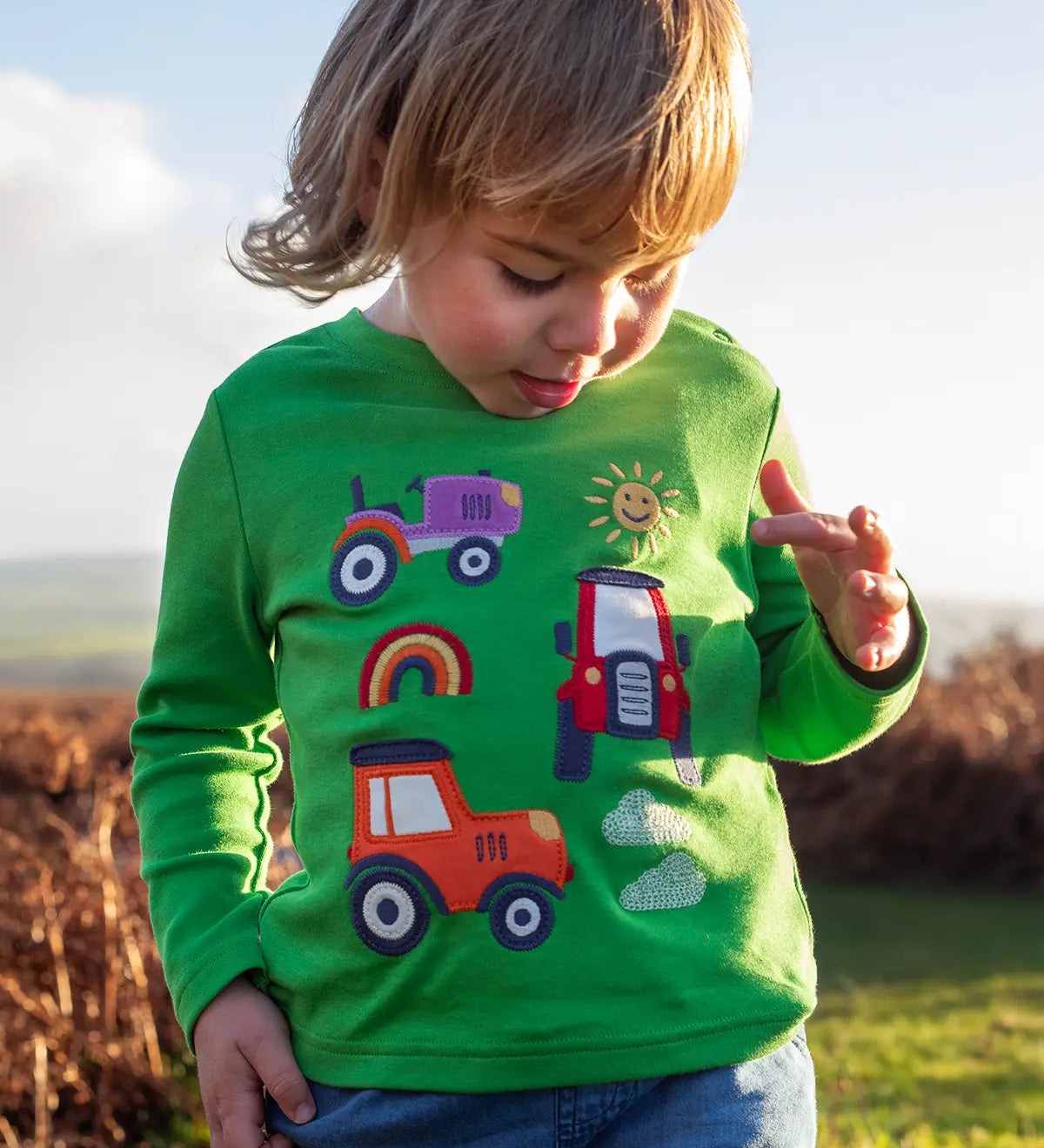 Front of child wearing Frugi's  GOTS organic cotton long sleeve top in green with playful tractor appliques 