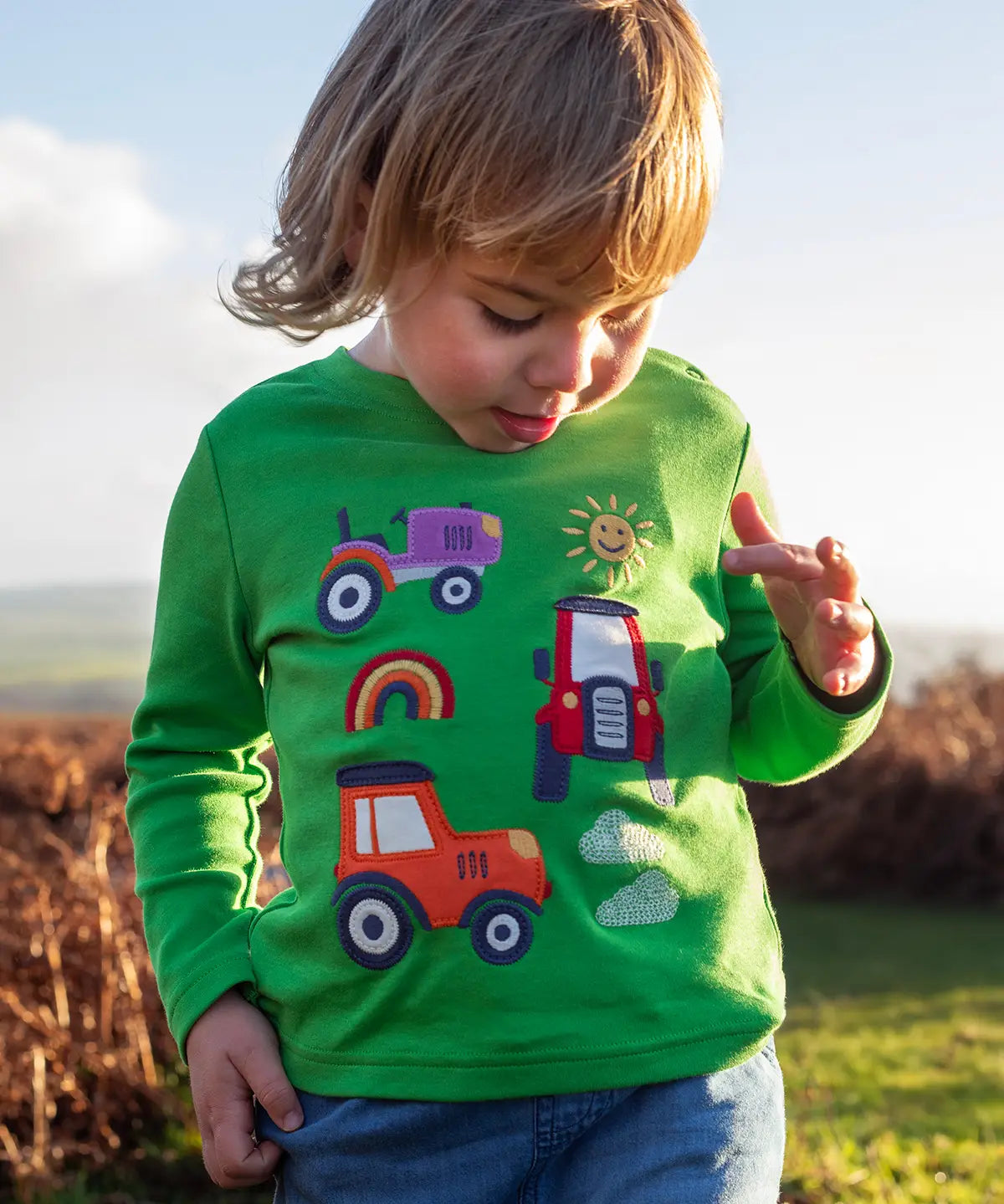Front of child wearing Frugi's  GOTS organic cotton long sleeve top in green with playful tractor appliques 