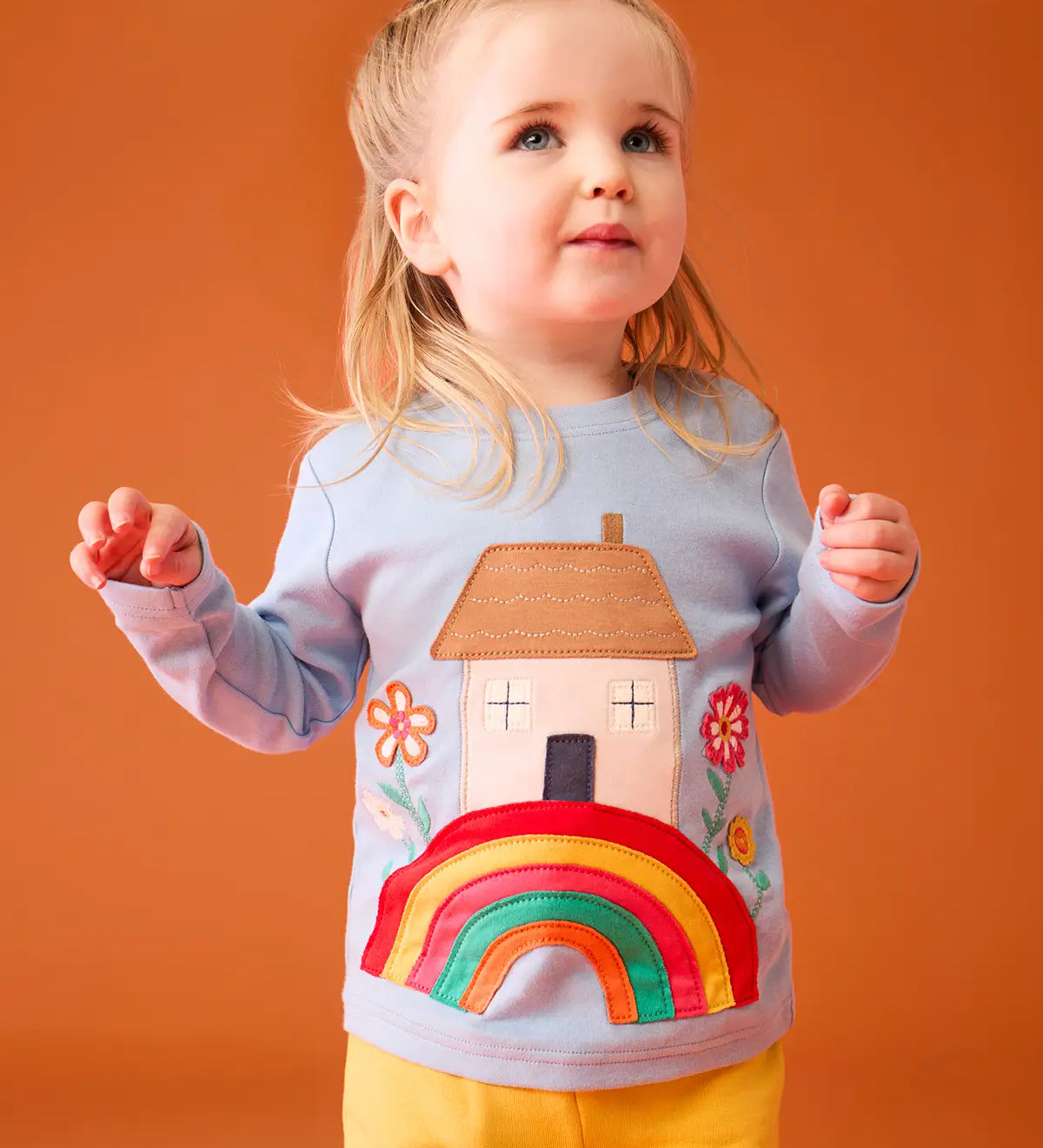 Small child wearing the Frugi's GOTS 100% organic cotton top in light blue showing the cute house on a rainbow applique on the front