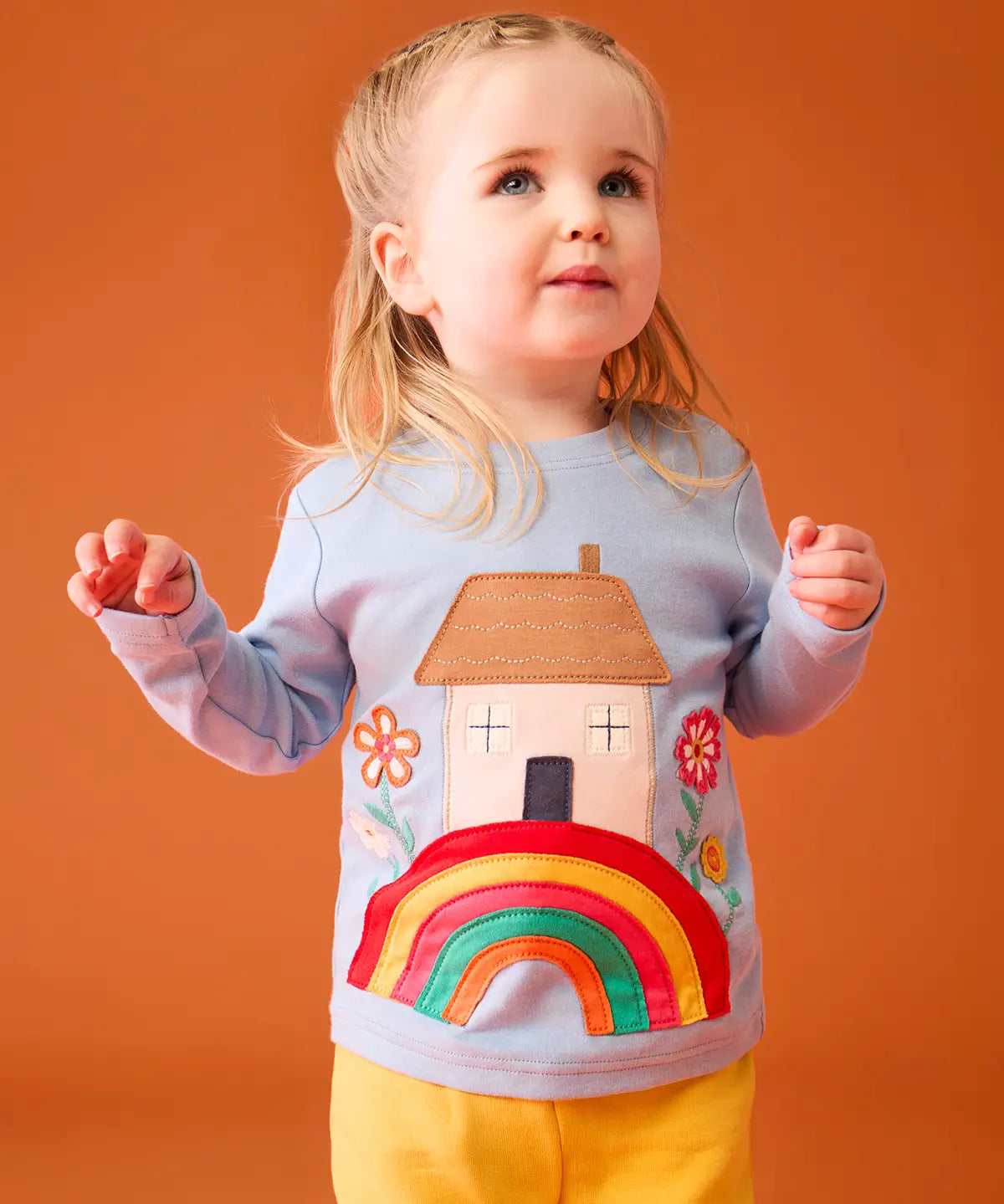 Small child wearing the Frugi's GOTS 100% organic cotton top in light blue showing the cute house on a rainbow applique on the front