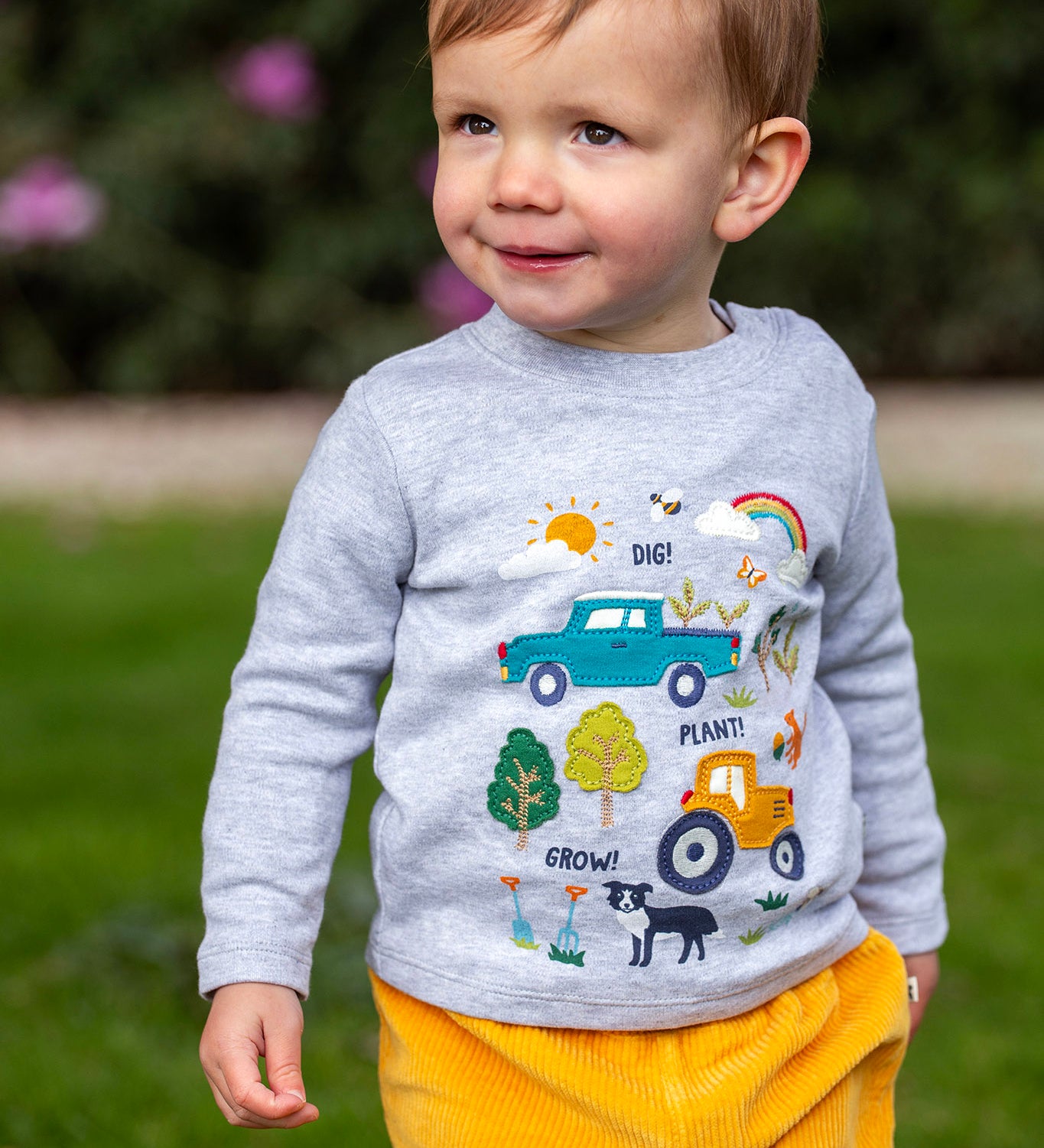 A toddler standing outside and wearing the Frugi kids adventure circular top in grey marl with farm inspired appliques on the front available at Babipur. 