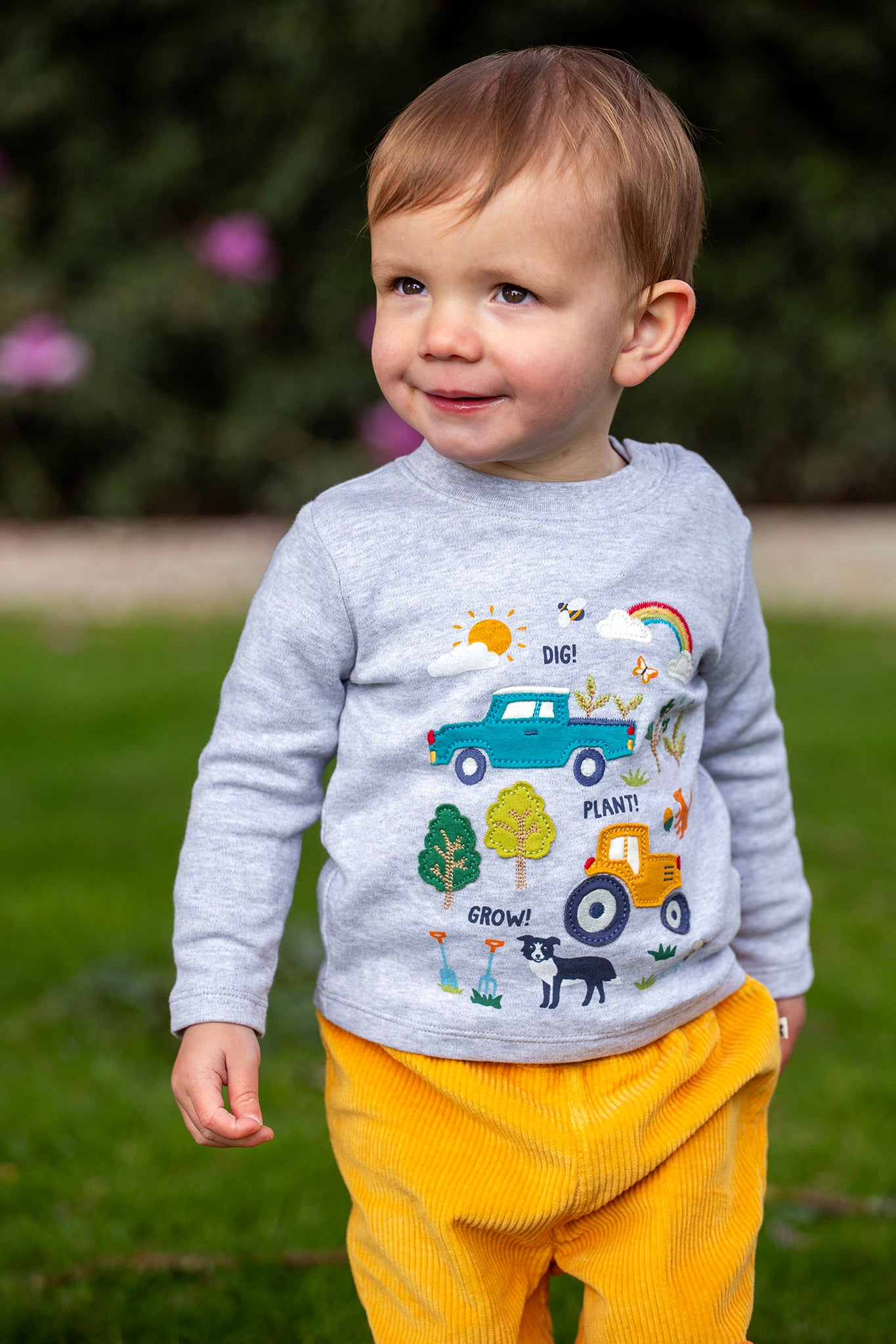 A toddler standing outside and wearing the Frugi kids adventure circular top in grey marl with farm inspired appliques on the front available at Babipur. 
