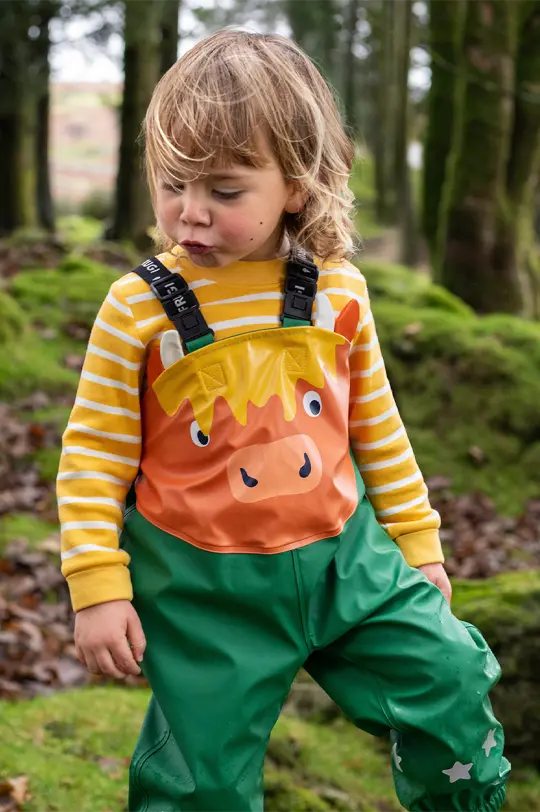 Highland Cow Frugi Puddle Buster pull up waterproof trousers worn by child with a striped yellow and white long sleeve top in organic cotton, in a forest.