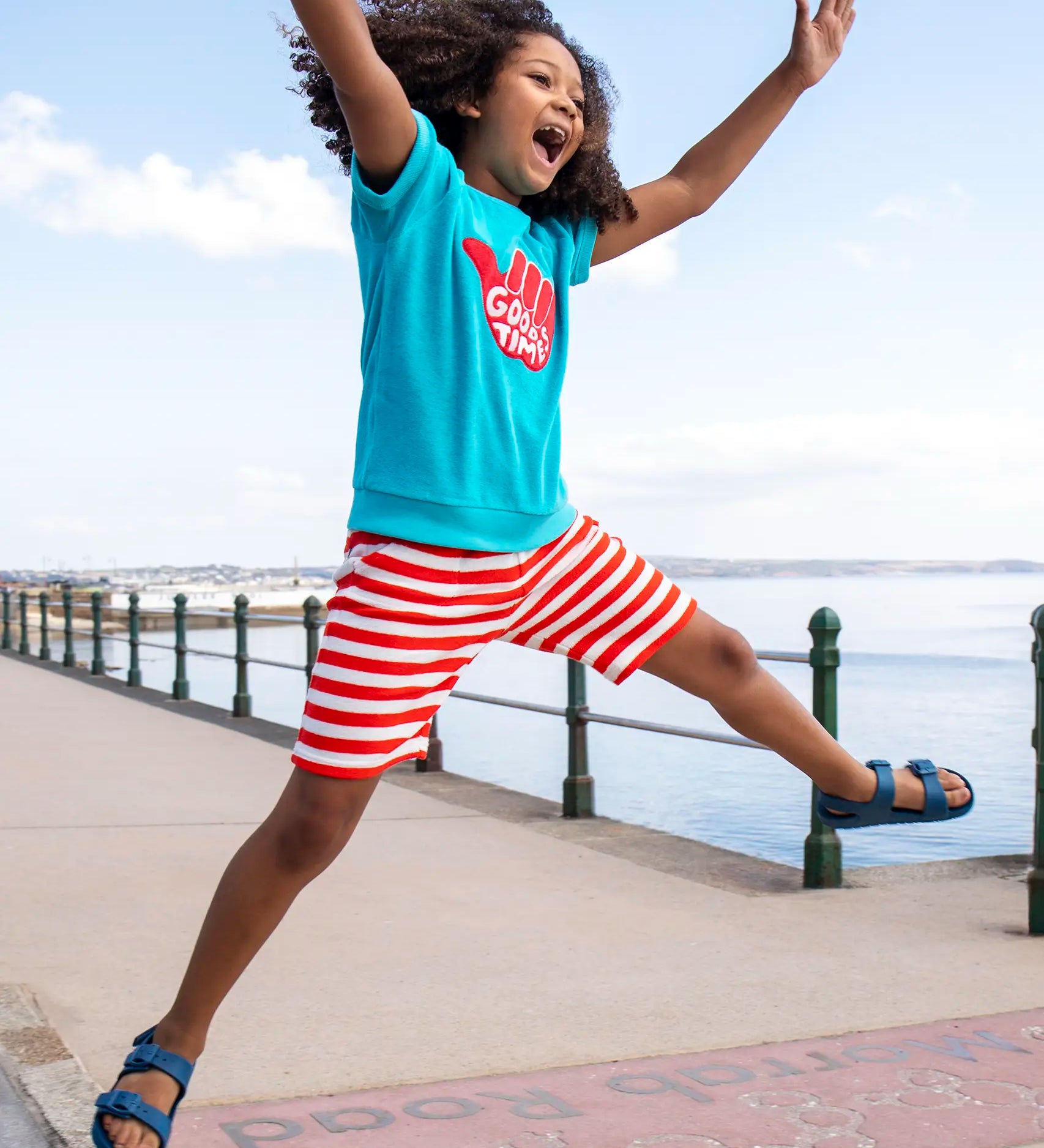 Child wearing Frugi's 100% organic cotton beach towelling shorts in red and white stripe and jumping at a beach promenade