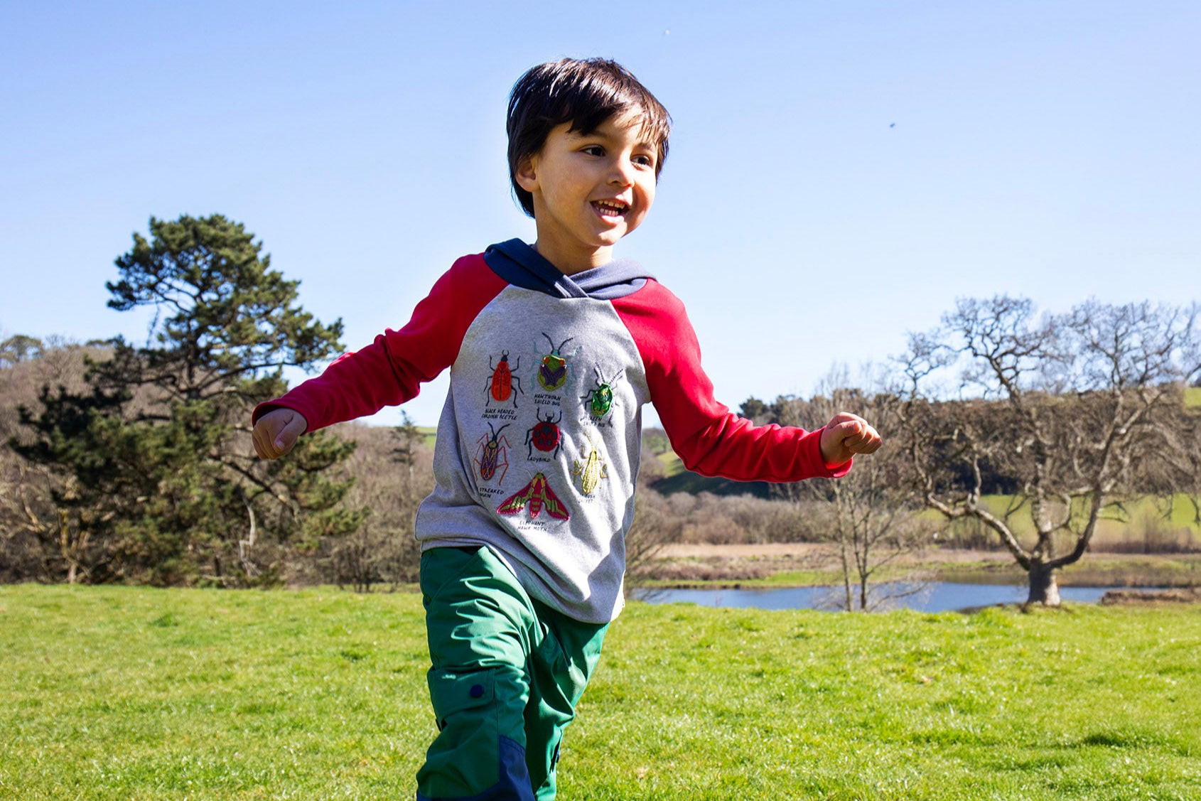 A child running outside on grass and wearing the Frugi compass kids hooded top in grey with red raglan sleeves and navy hood available at Babipur. 