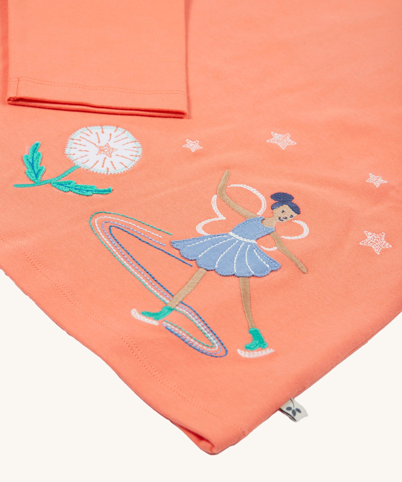 A closer look at the ice skating fairy on the Frugi Anna Applique Top - Cantaloupe/Fairy. The fairy wears a lilac dress with stars above and a dandelion to the side