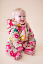 A toddler sitting down and wearing the Frugi kids cosy ted snuggle suit in mushroom field available at Babipur. A thick all in one suit in beige with a cute mushroom and flower design