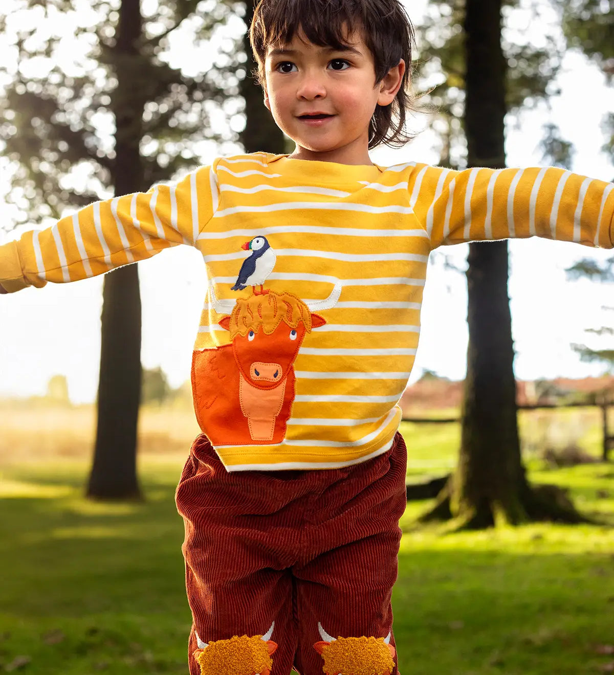 Child wearing Frugi's organic cotton kids long sleeve top in yellow with white stripes showing  highland cow with a puffin applique on the front