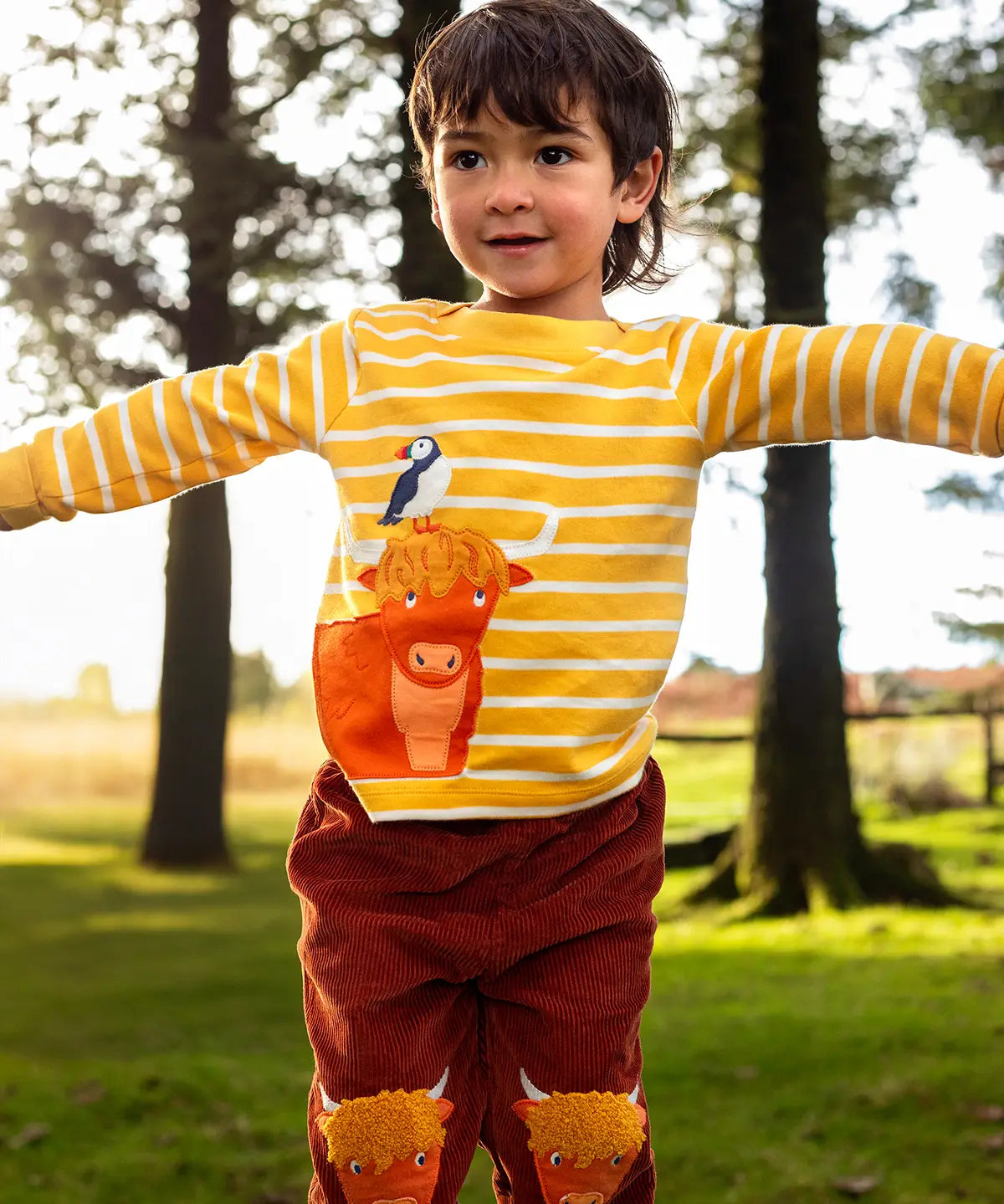 Child wearing Frugi's organic cotton kids long sleeve top in yellow with white stripes showing  highland cow with a puffin applique on the front