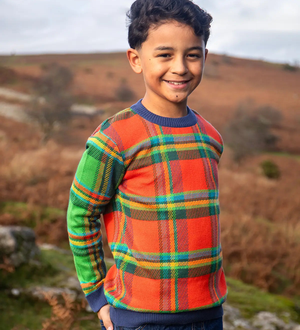 Child wearing Frugi's 100% GOTS organic cotton knitted kids jumper in a traditional tartan design outdoors. Jumper has a relaxed fit